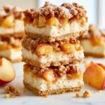 Peach cobbler cheesecake bars stacked with a buttery graham cracker crust, creamy cheesecake filling, juicy peaches, and cinnamon streusel topping.